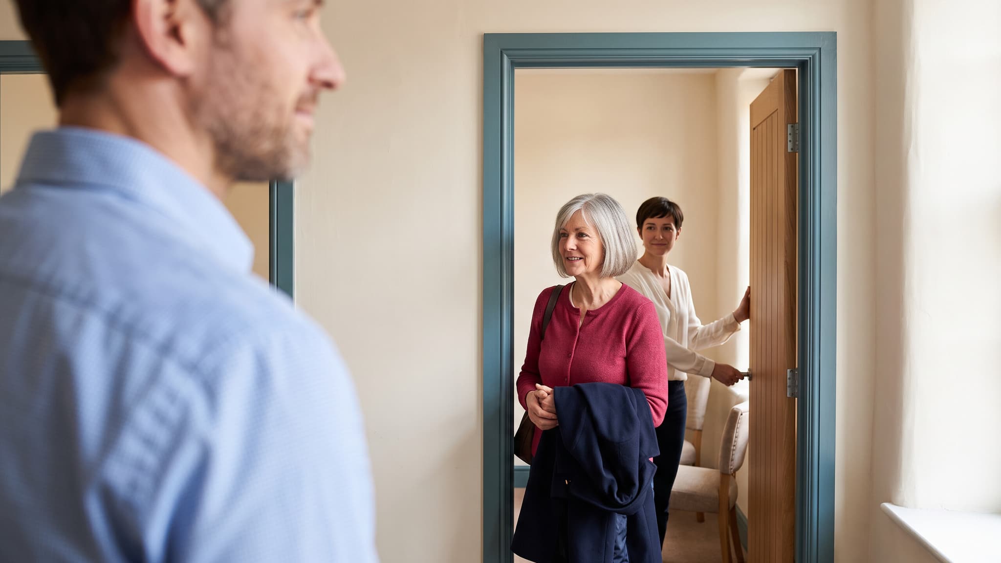 A practitioner waits as a patient leaves a shared consultation room and a colleague holds the door open.